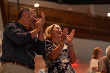 A couple in the audience enthusiastically applauds during a performance, smiling and clapping with joy. The warm lighting and blurred background capture the lively atmosphere of the event.