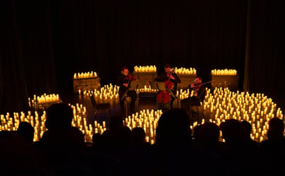 A string trio performs on a dimly lit stage surrounded by an array of glowing candles, creating an intimate and enchanting atmosphere. The audience watches from the foreground, silhouetted in shadows.